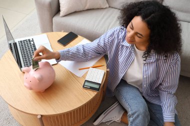 Laptop ve hesap makineli genç Afro-Amerikalı kadın kumbaraya dolar banknotu koyuyor.