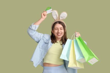 Yeşil arka planda kredi kartları ve alışveriş torbaları olan tavşan kulaklı genç bir kadın. Paskalya kutlaması