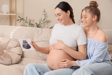 Evdeki koltukta oturan ultrason görüntüsü olan genç hamile lezbiyen çift.