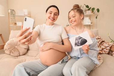 Sonogram görüntüsü olan genç hamile lezbiyen çift evdeki kanepede selfie çekiyor.
