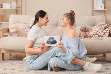 Evdeki halıda oturan ultrason görüntüsü olan genç hamile lezbiyen çift.