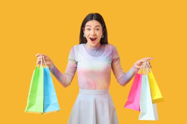 Sarı arka planda alışveriş torbaları olan şaşırmış genç bir kadın.