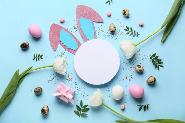 Boş karttan yapılmış çerçeve, kağıttan tavşan kulaklar, paskalya yumurtaları ve mavi arka planda lale çiçekleri.