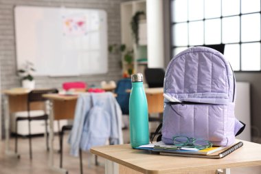 Sınıftaki masasında gözlük ve su şişesi olan leylak rengi sırt çantası.