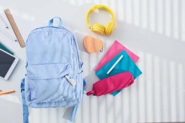 Okul kırtasiyesi ve kulaklıklı sırt çantası beyaz masada, üst görünümde.