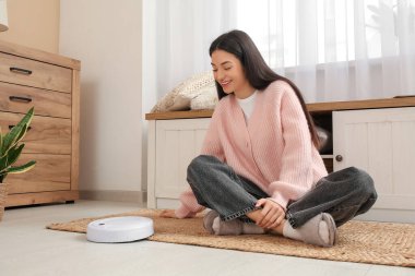 Evde halıya oturmuş robot süpürgesi olan genç bir kadın.