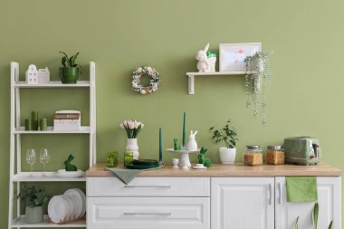 Mutfağın içinde raflar, Paskalya çelengi, çiçekler ve çatal bıçaklar yeşil duvarın yanında.