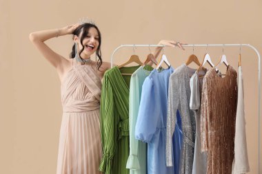 Güzel, mutlu genç kız tacın yanında. Bej arka planda şık balo elbiseleri var.