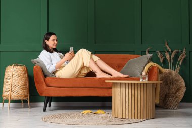 Evdeki yeşil duvarın yanındaki kırmızı koltukta cep telefonu kullanan genç bir kadın.