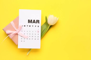 Sarı arka planda takvim, lale çiçeği ve Kadınlar Günü kutlaması için hediye paketi.