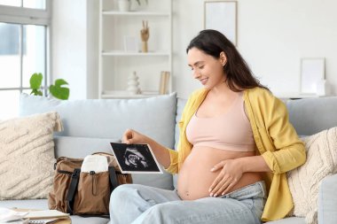 Evdeki kanepede oturan ultrason görüntüsü ve doğum çantası olan hamile bir kadın.