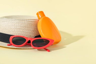 Güneş gözlüklü bir şişe güneş kremi ve açık sarı arka planda yaz şapkası.