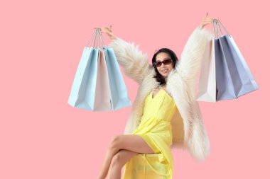 Güneş gözlüklü, sandalyede pembe arka planda oturan mutlu ve güzel Asyalı kadın.