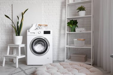 Çamaşır makinesi, raf ünitesi ve ev bitkileriyle dolu rahat bir çamaşırhane.