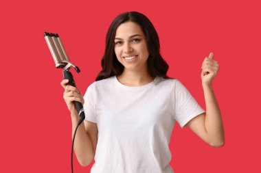 Kırmızı arka planda baş parmağını gösteren saç maşası olan güzel genç bir kadın.