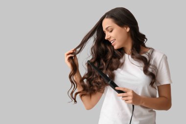 Beautiful young woman with curling iron on grey background