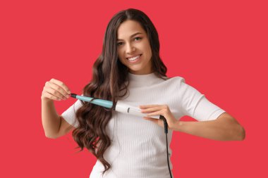Beautiful young woman with curling iron on red background