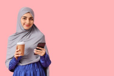 Pembe arka planda bir fincan kahve ve cep telefonu olan güzel Müslüman kadın.