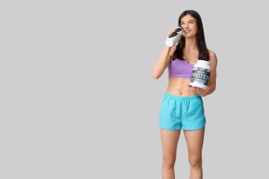 Gri arka planda protein tozu içen genç sporcu kadın.