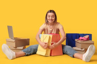 Alışveriş torbaları ve dizüstü bilgisayarlı genç bir kadın sarı arka planda oturuyor.