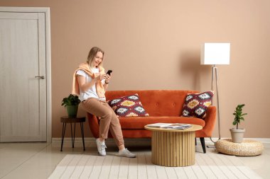 Genç ve güzel bir kadın cep telefonu kullanıyor ve bej oturma odasında kanepede oturuyor.