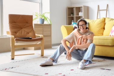 Gözlüklü ve kulaklıklı genç bir kadın evde yerde müzik dinliyor.