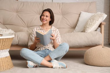 Genç, güzel bir kadın kitap okuyor ve oturma odasındaki kanepenin yanında oturuyor.