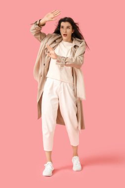 Young woman in trench coat with scarf and strong wind on pink background