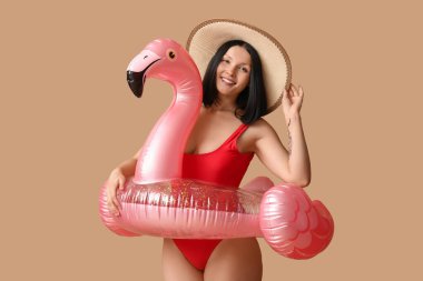 Havalı kırmızı mayo giyen, kahverengi arka planda flamingo şeklinde şişirilebilir yüzük takan güzel bir kadın.