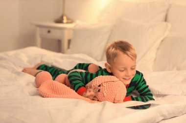 Oyuncak tavşancığıyla küçük şirin bir çocuk gece yatak odasında tablet bilgisayardan çizgi film izliyor.