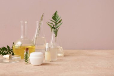 Pembe duvarın yanındaki bej masada doğal kozmetik ürünleri hazırlamak için malzemeler ve numunelerle dolu laboratuvar vitrinleri.