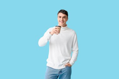 Süslü beyaz süveter içinde, mavi arka planda bir fincan sıcak kahveyle yakışıklı bir genç.