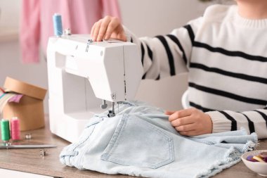 Genç bayan terzi atölyenin yanındaki masanın yanında kot şort dikmek için dikiş makinesi kullanıyor.