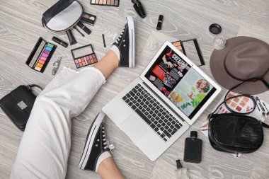 Modern dizüstü bilgisayarlı kadın bacakları, farklı dekoratif kozmetik ürünleri ve hafif ahşap zeminde şık aksesuarlar. Çevrimiçi alışveriş kavramı