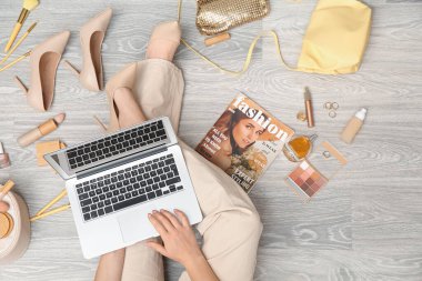 Modern dizüstü bilgisayarı, farklı dekoratif kozmetik ürünleri, aksesuarları ve dergisi olan bir kadın açık tahta zeminde oturuyor. Çevrimiçi alışveriş kavramı