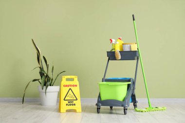 Trolley 'de temizlik malzemeleri ve yeşil duvarın yanında uyarı levhası var.