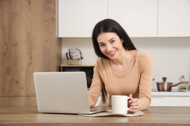 Mutfakta dizüstü bilgisayar kullanarak çay içen güzel genç bir kadın.