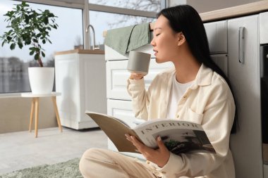 Genç Asyalı kadın mutfakta kahve içiyor ve dergi okuyor.