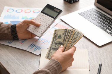 Ofiste hesap makinesi kullanırken erkek eller dolar banknotlarını sayıyor. Yakın çekim.