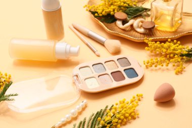 Süslü aksesuarları olan dekoratif kozmetik ürünleri ve turuncu arka planda mimoza çiçekleri, yakın plan.