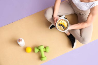 Spor aletleri olan olgun bir kadın, bir kase besleyici yulaf ezmesi ve yoga minderinde oturan meyveler, üst manzara.