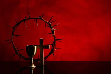 Dikenli taç, haç ve şarap kadehi siyah masada, kırmızı arka planda.