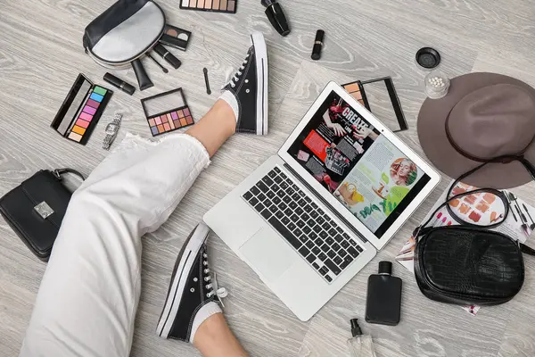 Modern dizüstü bilgisayarlı kadın bacakları, farklı dekoratif kozmetik ürünleri ve hafif ahşap zeminde şık aksesuarlar. Çevrimiçi alışveriş kavramı