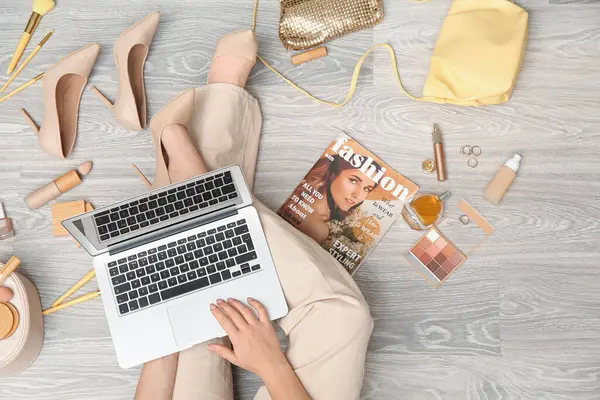 Modern dizüstü bilgisayarı, farklı dekoratif kozmetik ürünleri, aksesuarları ve dergisi olan bir kadın açık tahta zeminde oturuyor. Çevrimiçi alışveriş kavramı