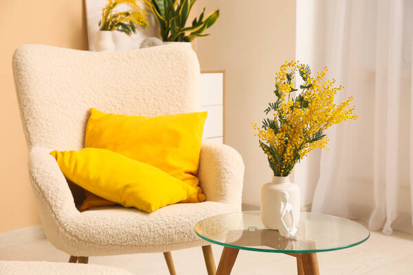 Vase with mimosa flowers and statuette on table near armchair in living room