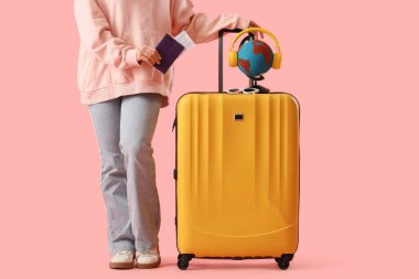 Pasaportu ve bavulunda küresi olan kadın turist pembe arka planda.