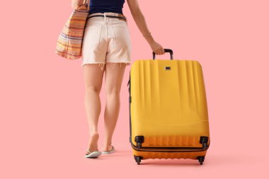 Plaj çantalı ve bavullu bir kadın turist pembe arka planda yürüyor.