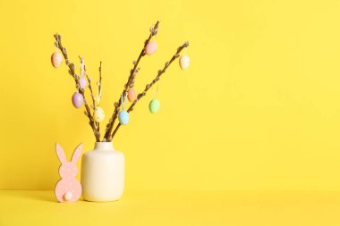 Sarı arka planda söğüt dalları ve renkli Paskalya yumurtaları olan vazo.