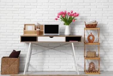 Modern dizüstü bilgisayarlı şık çalışma alanı, bir buket güzel lale çiçeği ve beyaz tuğla duvarın yanındaki raf ünitesi.