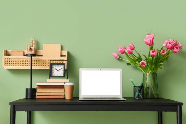 Modern dizüstü bilgisayarı, kitapları ve yeşil duvarın yanındaki güzel lale çiçekleriyle süslü çalışma alanı.